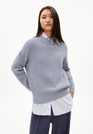 Hellgrauer Strickpullover mit gerippter Textur und hohem Kragen, über einem weißen Hemd getragen, kombiniert mit dunklen Hosen.