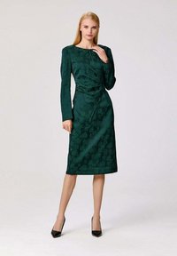 Robe verte à motifs avec des manches longues, présentant une silhouette ajustée et un détail drapé sur le devant, associée à des talons hauts noirs.