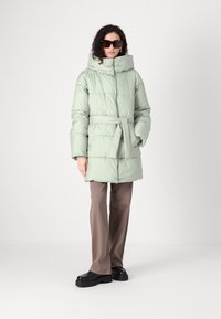 Lys grønn dunjakke med belte i livet, høy krage og horisontalt quiltet design. Kombinert med brune bukser og svarte klumpete sko.