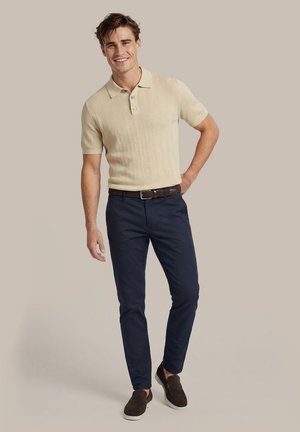 Junger Mann lächelt, trägt beige kurzärmeliges Polo, navy Slim-Fit-Hose mit braunem Gürtel und braune Slipper, steht auf neutralem Hintergrund.