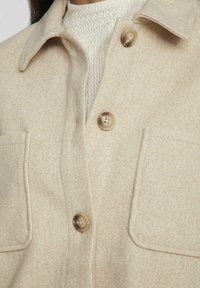 Giacca camicia in misto lana beige con colletto, due tasche a toppa e grandi bottoni marroni. La texture è morbida e leggermente strutturata.