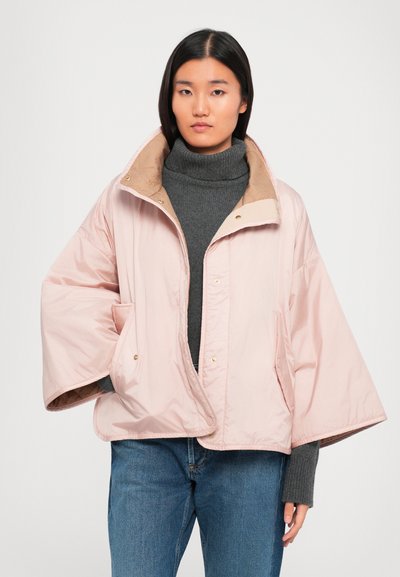 Mladá žena v svetloružovej oversized jasnéj bunde, pod ktorou má na sebe šedý rolák a modré džínsy, stojí na jednoliatom pozadí.