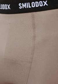 Nahaufnahme eines beigen Stoffes mit vertikaler Naht und schwarzem Bund, auf dem weißer Text „SMILODOX" zu sehen ist.