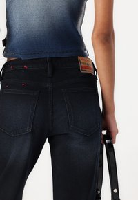 Jean en denim foncé avec taille haute, présentant des accents de surpiqûres rouges et un patch en cuir. Les poches arrière sont bien visibles.