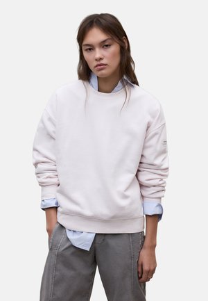 Giovane donna con capelli castani indossa una felpa rosa chiaro sopra una camicia blu, abbinata a pantaloni grigi, in piedi con le mani nelle tasche.