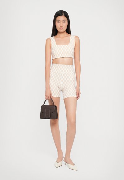 Two-piece outfit in light cream with geometric patterns; cropped square top and high-waisted shorts. Brown handbag and white heeled mules.