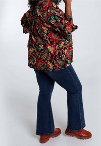 Blusa a fiori con maniche corte in colori vivaci (rosso, verde, arancione) abbinata a jeans a zampa d'elefante blu scuro e scarpe spesse marroni.