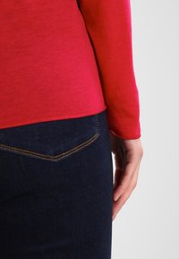 Rotes Langarmoberteil mit glatter Textur, kombiniert mit dunkelblauen Jeans, die auffällige Nähte und ein Detail auf der Hintertasche aufweisen.