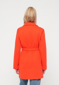 Casaco vermelho com cintura cingida, mangas longas e um colarinho largo. Tecido suave e texturizado, design até aos joelhos. Vista traseira mostrada.