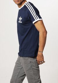 Námořnická modrá bavlněná tričko s bílými sportovními pruhy na rukávech, s kontrastním bílým logem Adidas na hrudi. Krátké rukávy, kulatý výstřih.