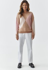 Maglione lavorato a maglia con un motivo geometrico nei colori rosa chiaro, beige e crema; abbinato a pantaloni bianchi larghi e scarpe nere.
