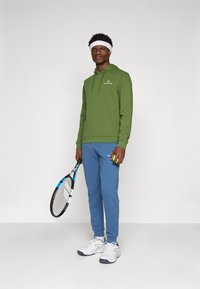 Grønn hettegenser med kengurulomme, blå joggebukser og hvite joggesko. Holder en tenniskølle og en grønn tennisball.