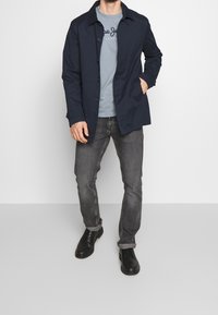 Marineblauwe jas met knoopsluiting, lichtblauw t-shirt met logo, grijze jeans en zwarte schoenen. De jas heeft een kraag en een zijzak.