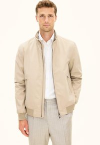 Ung mand med kort lyst brunt hår iført beige lynlåstrøje, hvid skjorte med striber og lysegrå bukser med nålestriber.