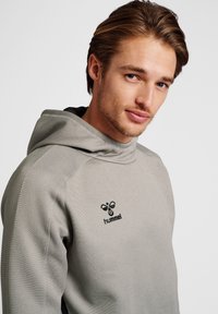 Sweatshirt à capuche gris en tissu texturé, arborant un logo noir sur la poitrine et des accents noirs sur les manches.
