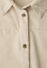 Devant d'une chemise en velours côtelé beige avec col et deux boutons marron alignés verticalement sur la patte de boutonnage.
