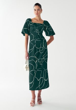 Dunkelgrünes Midi-Kleid mit Puffärmeln und quadratischem Ausschnitt. Hat ein weißes, abstraktes Blumenmuster. Das Modell hält eine kleine weiße Handtasche.