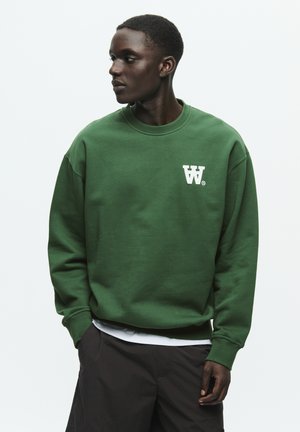 Green sweatshirt made of cotton, featuring a crew neckline and embroidered logo on the left chest. Model paired with dark shorts.