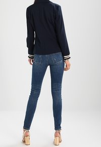 Granatowa marynarka o dopasowanym kroju z białymi paskami przy mankietach, w połączeniu z dżinsami w slim fit w odcieniu niebieskim. Sandały na obcasie o masywnej konstrukcji.