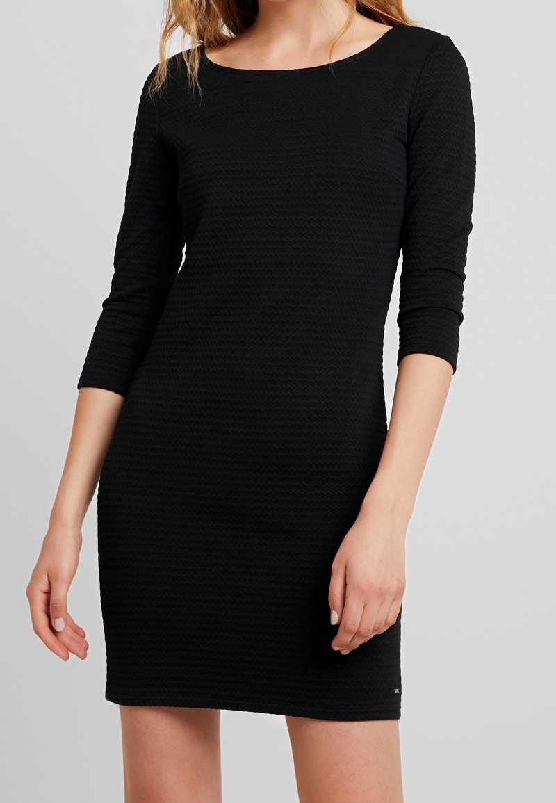 Musta, vartalonmyötäinen mekko, joka on valmistettu rakenteellisesta kankaasta, jossa on venepääntie ja kolmen neljäsosan hihat. Ominaisuuksiin kuuluu yksinkertainen muotoilu ilman kuvioita tai varusteita.