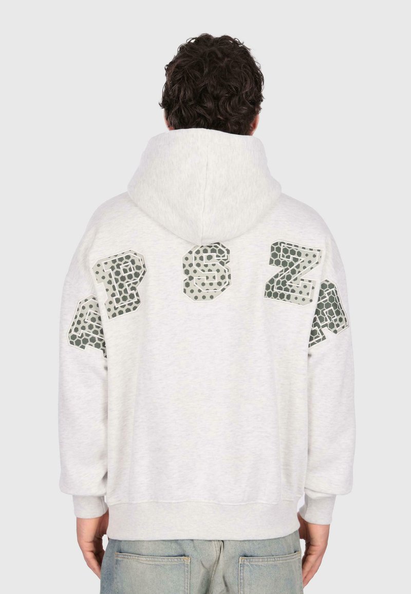 Homme portant un sweat à capuche gris clair avec de grandes lettres à motifs dans le haut du dos et un jean bleu clair, tourné le dos à l'appareil photo.