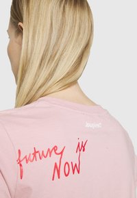 Una persona con pelo largo y rubio se ve de espaldas, llevando una camisa rosa con el texto "el futuro es ahora" en cursiva roja en la parte posterior.