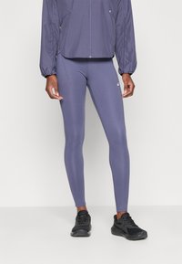 Paarse, strakke legging gemaakt van rekbaar materiaal, gecombineerd met een bijpassende jas met elastische mouwen en een ritssluiting aan de voorkant. Zwarte sportieve schoenen.