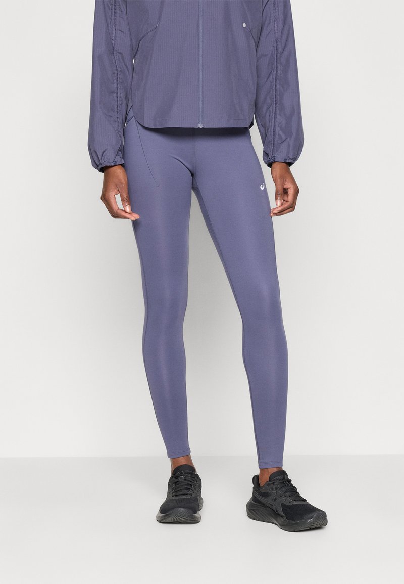 Paarse, strakke legging gemaakt van rekbaar materiaal, gecombineerd met een bijpassende jas met elastische mouwen en een ritssluiting aan de voorkant. Zwarte sportieve schoenen.