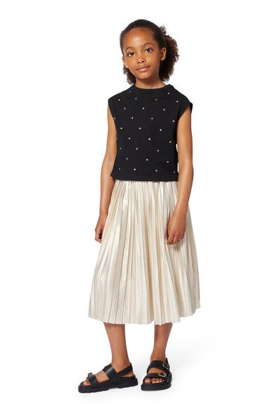 Haut sans manches noir avec des clous dorés, associé à une jupe plissée de couleur crème. Des sandales noires avec un détail de boucle complètent la tenue.