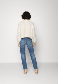 Suéter branco de malha trançada com ombros caídos, combinado com jeans de perna reta azul claro e sapatos de salto brancos. Vista traseira, fundo minimalista.