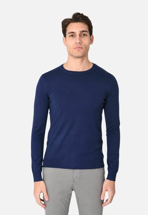 Uomo adulto con maglione aderente a maniche lunghe blu navy e pantaloni grigio chiaro, in piedi davanti a uno sfondo bianco semplice.