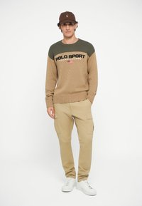 Jersey de punto en color caqui y verde oliva con un patrón texturizado, que presenta el logo "POLO SPORT". Combinado con pantalones cargo beige y zapatillas blancas.