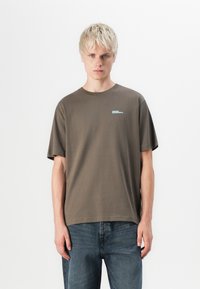 Bruine katoenen t-shirt met korte mouwen, ronde halslijn en een klein blauw logo op de borst. Gecombineerd met blauwe spijkerbroek.