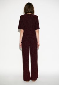 Bordeaux jumpsuit van een gestructureerde glinsterende stof. Het heeft korte mouwen, een nauwsluitende taille, uitlopende benen en een ritssluiting aan de achterzijde.