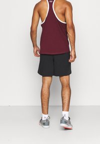 Bordowy bezrękawnik z białymi wykończeniami, w połączeniu z czarnymi szortami i szarymi butami sportowymi. Skarpetki mają wzór, logo widoczne na odzieży.