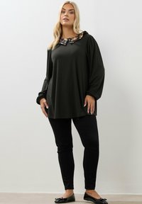 Donna in piedi che indossa una tunica scura a maniche lunghe con colletto floreale, pantaloni neri e ballerine nere su uno sfondo chiaro e semplice.