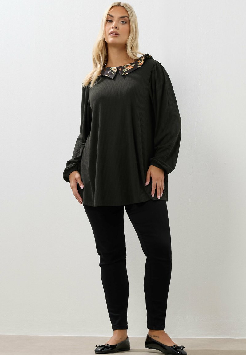 Donna in piedi che indossa una tunica scura a maniche lunghe con colletto floreale, pantaloni neri e ballerine nere su uno sfondo chiaro e semplice.