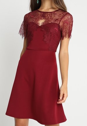 Woman wearing a knee-length, burgundy dress with lace short sleeves and lace detail on the upper bodice and neckline.