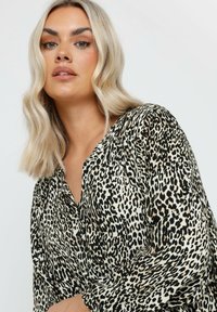 Blusa con stampa leopardata realizzata in tessuto leggero, con colletto e chiusura a bottone. Il motivo è composto da macchie nere e marroni su uno sfondo crema.