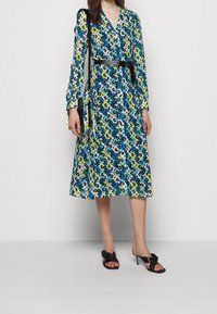 Robe portefeuille florale en bleu, vert et noir avec des manches longues, un col en V, une taille ceinturée et une jupe fluide. Associée à des sandales à talons noires.