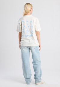 Crème-kleurig katoenen t-shirt met een blauwe afbeelding van negen aardbeien en de tekst "TASTE OF SUMMER" op de achterkant. Ontspannen pasvorm, korte mouwen.
