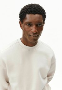 Felpa beige chiaro con scollo rotondo e dettagli a costine sui polsini, con una texture liscia. Primo piano del volto di una persona, vista frontale.