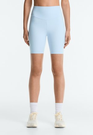Personne portant un short de cyclisme bleu clair taille haute, des chaussettes blanches et des baskets beiges, debout sur un fond blanc.