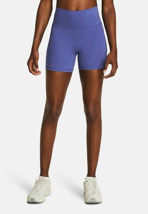 Atleet draagt blauw compressieshort met hoge taille en witte hardloopschoenen, staat met iets gespreide benen op een witte achtergrond.