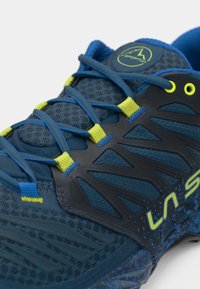 Μπλε αθλητικό παπούτσι με κλειστό πλέγμα, μαύρες λεπτομέρειες, φωτεινά κίτρινα κορδόνια και προστατευτικό καπάκι από καουτσούκ στην μύτη.