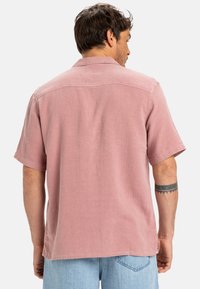 Koszula z krótkim rękawem w jasnoróżowym lnie, o luźnym kroju, z otwartym kołnierzykiem i rozcięciami po bokach. Z tyłu znajduje się prosty detal szwu.