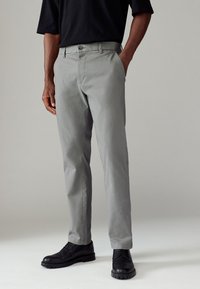 Pantalons gris avec une finition texturée, design ajusté, et fermeture à bouton. Portés avec des chaussures de ville noires, illustrant un look décontracté.