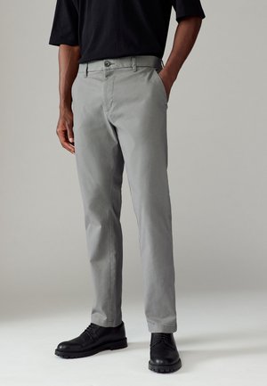 Pantalons gris avec une finition texturée, design ajusté, et fermeture à bouton. Portés avec des chaussures de ville noires, illustrant un look décontracté.
