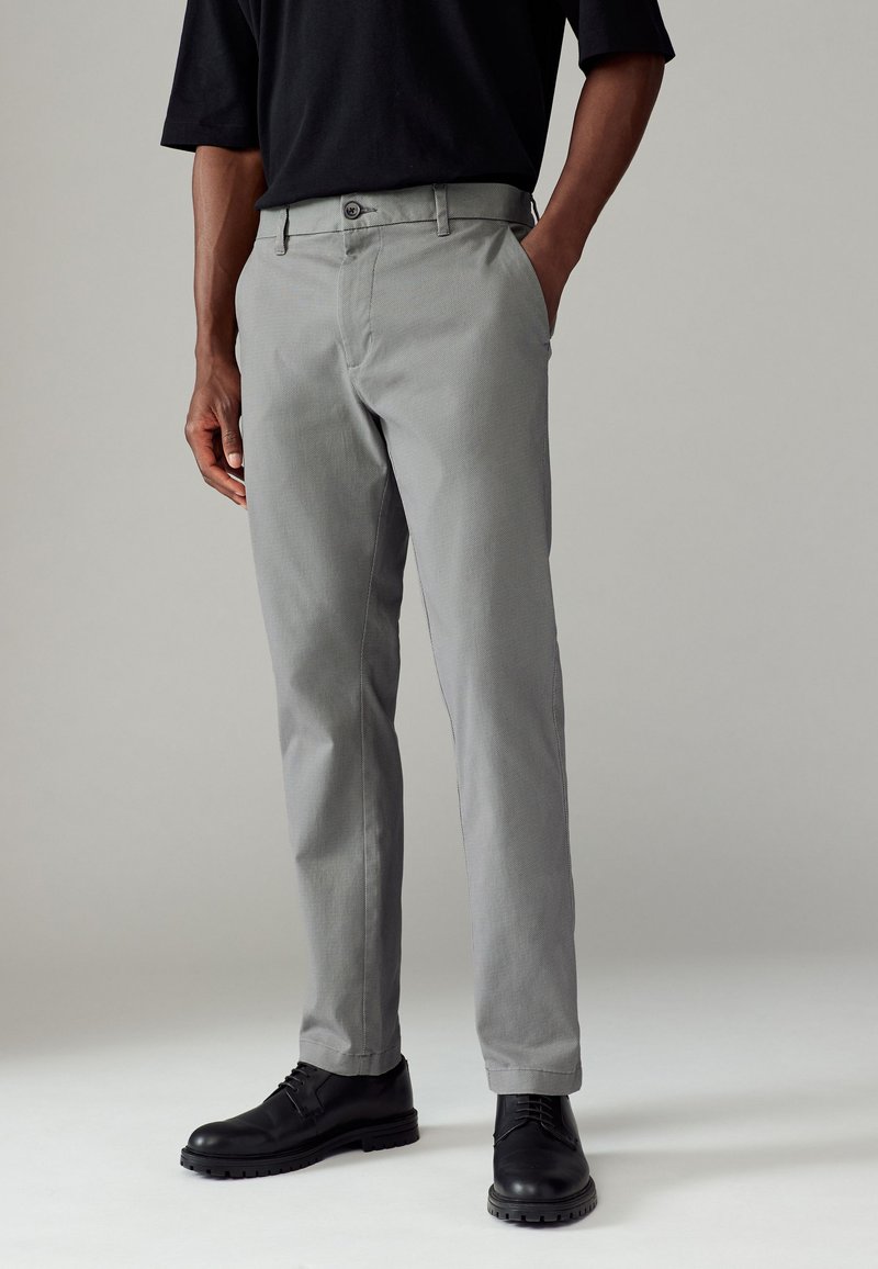 Pantalons gris avec une finition texturée, design ajusté, et fermeture à bouton. Portés avec des chaussures de ville noires, illustrant un look décontracté.