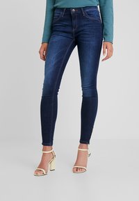 Jeans ajustados de denim azul oscuro con un leve desgastado, diseño de cinco bolsillos y cintura media. Combinados con sandalias de tacón doradas.
